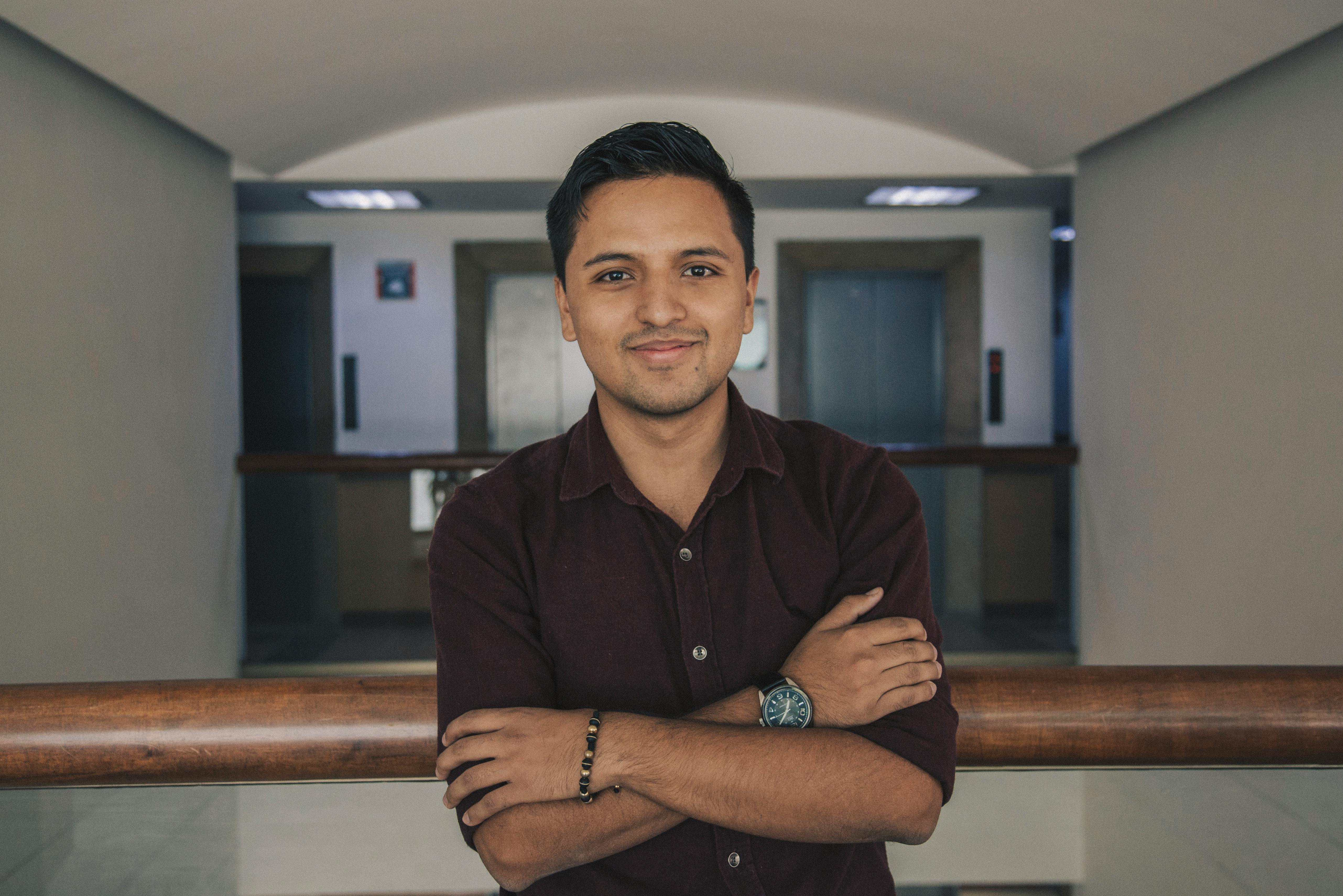 Positive Indian in casual outfit standing with crossed arms near glass fence while smiling and looking at camera in modern corridor